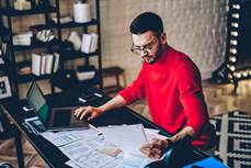 Concentrated young man in eyeglasses reading information from paper documents and checking in database...